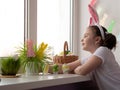 A little girl with rabbit ears in a white sweater stands hopefully looking out the window with her head tilted. Concept Easter