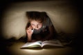 Little girl is reading a book under a blanket with a flashlight in a dark room at night Royalty Free Stock Photo