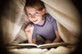 Little girl is reading a book under a blanket with a flashlight in a dark room at night Royalty Free Stock Photo