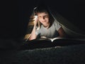 little girl is reading a book under a blanket with a flashlight in a dark room at night Royalty Free Stock Photo