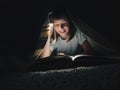 little girl is reading a book under a blanket with a flashlight in a dark room at night Royalty Free Stock Photo