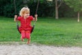 Little girl in red dress sitting on swing. Happy smiling child swinging in park. Kid at playground. Royalty Free Stock Photo