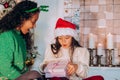 Little girl in a red hat opens a Christmas present from mom against the background of a Christmas tree and candles Royalty Free Stock Photo
