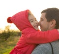 A little girl in a red jacket with a hood hugs and kisses her dad, smiles, touches her nose. Happy family, father`s day, bright ra