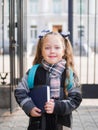 A little girl with red tails in her hands is holding a book in front of the school.