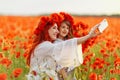 Little girl with redhead mother in white dresses and wreathes make selfie with bouquet of poppies on poppy field at summer sunset