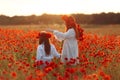 Little girl with redhead mother in white dresses and wreathes walking on poppy field with bouquet of poppies at summer sunset Royalty Free Stock Photo