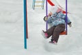 Little girl is riding on frosty winter playgrounds swing covering with white snow outdoors