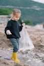 Little girl in rubber boots with a bag walks through shallow water looking for garbage Royalty Free Stock Photo