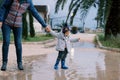 Little girl in rubber boots stands in a puddle holding her mother hand. Cropped