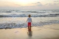 A little girl on a sandy beach against the background of the setting sun went into the sea and looks into the distance