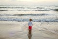 A little girl on a sandy beach against the background of the setting sun went into the sea and looks into the distance