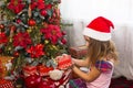 Little girl in Santa hat opens a red box with a gift and a Golden magic light near the Christmas tree. Holiday decor, poinsettias Royalty Free Stock Photo