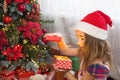 Little girl in Santa hat opens a red box with a gift and a Golden magic light near the Christmas tree. Holiday decor, poinsettias Royalty Free Stock Photo