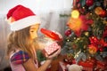 Little girl in Santa hat opens a red box with a gift and a Golden magic light near the Christmas tree. Holiday decor, poinsettias Royalty Free Stock Photo