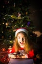 Little girl in a Santa hat and red dress under Christmas tree is dreaming, waiting for the holiday, lying on a plaid blanket. A Royalty Free Stock Photo