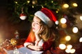 Little girl in a Santa hat and red dress under Christmas tree is dreaming, waiting for the holiday, lying on a plaid blanket. A Royalty Free Stock Photo