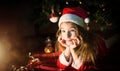 Little girl in a Santa hat and red dress under Christmas tree is dreaming, waiting for the holiday, lying on a plaid blanket. A Royalty Free Stock Photo