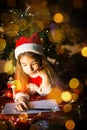 Little girl in a Santa hat and red dress under Christmas tree is dreaming, waiting for the holiday, lying on a plaid blanket. A Royalty Free Stock Photo