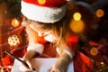 Little girl in a Santa hat and red dress under Christmas tree is dreaming, waiting for the holiday, lying on a plaid blanket. A Royalty Free Stock Photo