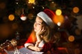 Little girl in a Santa hat and red dress under Christmas tree is dreaming, waiting for the holiday, lying on a plaid blanket. A Royalty Free Stock Photo