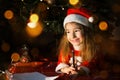 Little girl in a Santa hat and red dress under Christmas tree is dreaming, waiting for the holiday, lying on a plaid blanket. A Royalty Free Stock Photo