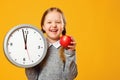 Little girl schoolgirl holds a big clock and a red apple on a yellow background. Break time and lunch