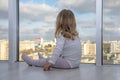 A little girl sits on the floor of an apartment in front of huge Windows and looks down at the city. Concept: children`s lonelines