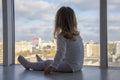 A little girl sits on the floor of an apartment in front of huge Windows and looks down at the city. Concept: children`s lonelines