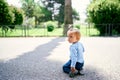 Little girl sits on her knees on the asphalt against the background of a fence in a green park Royalty Free Stock Photo