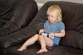 A little girl sits on a leather sofa and controls a game joystick in her hands and plays a console Royalty Free Stock Photo