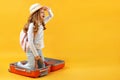 A little girl sits in a suitcase and looks into the distance on a yellow background. The child dreams of travel, trip, adventure.