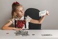 A little girl sits at a table in front of a stack of hundred-dollar bills and examines one of them for light, checks for falsity Royalty Free Stock Photo