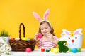 A little girl sits at a table and paints Easter eggs with a brush. Happy child in the ears of a bunny. Royalty Free Stock Photo