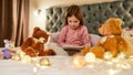 A little girl is sitting alone cross-legged barefoot on a huge bed reading a book enjoying her time with teddybears and Royalty Free Stock Photo