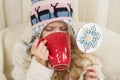 Little girl sitting at home in the chair, in knitted hat with Christmas ornament with cup of hot drink. Girl holding symbolic Royalty Free Stock Photo