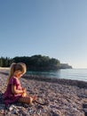 Little girl is sitting on a pebble beach by the sea and opening a small bag