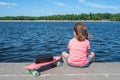 A little girl is sitting on a river bank with a music speaker and skateboard at the blue river background. A back view. Lifestyle