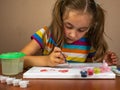 The little girl sitting at the table enthusiastically draws with a brush and watercolor paints on a white canvas of Royalty Free Stock Photo