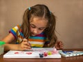 The little girl sitting at the table enthusiastically draws with a brush and watercolor paints on a white canvas of Royalty Free Stock Photo