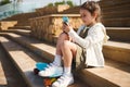 Little girl with skateboard, sitting on steps outdoors, using smartphone, checks social media content, watches cartoons