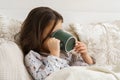 Little girl in sleepwear lie under blanket and take sip of drink covering face with mug. Unhealthy child, side view. Royalty Free Stock Photo