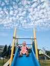 A little girl is sliding down a children`s slide against the background of a blue sky with white clouds. Royalty Free Stock Photo