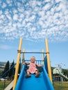 A little girl is sliding down a children's slide against the background of a blue sky with white clouds.