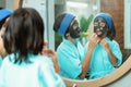 a little girl smiles while cleaning a black clay face mask by her mother in front of the mirror Royalty Free Stock Photo