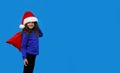 Little girl with smling wearing santa hat carrying gift bag isolated on blue background. Winter holidays. Copy space Royalty Free Stock Photo