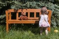 Little girl standing by the wooden bench with a teddy bear. Playing in the garden under a blue spruce. Sunny day in a garden. Royalty Free Stock Photo