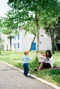 Little girl stands in front of mom and child sitting on the lawn under a tree Royalty Free Stock Photo