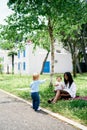 Little girl stands and points a finger at mom and child sitting on the lawn under a tree Royalty Free Stock Photo