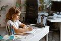 Little girl studies distantly at videocall via laptop at table in light room Royalty Free Stock Photo